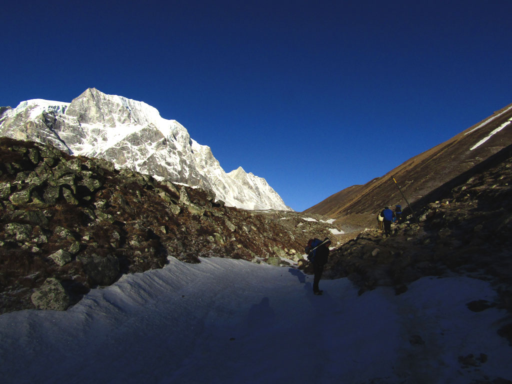 A porter pauses as the sunrise reaches the glacier trail from Larkye Phedi to Larkye La. Most trekkers begin the day's walk around 4am and walk for a couple of hours in freezing temperatures. At these altitudes is best to take things slow and reach the crest of the pass before mid-day. To the left are the peaks of the Larkye Himal